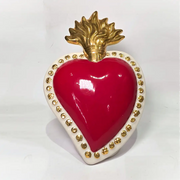 Scultura a forma di Cuore Sacro in ceramica rossa lucida, completata da una fiamma superiore dorata e una cornice bianca perlinata con borchie oro. Questo oggetto di design artigianale con base d'appoggio è un complemento d'arredo iconico, ideale per donare un tocco di eleganza e spiritualità moderna agli interni.