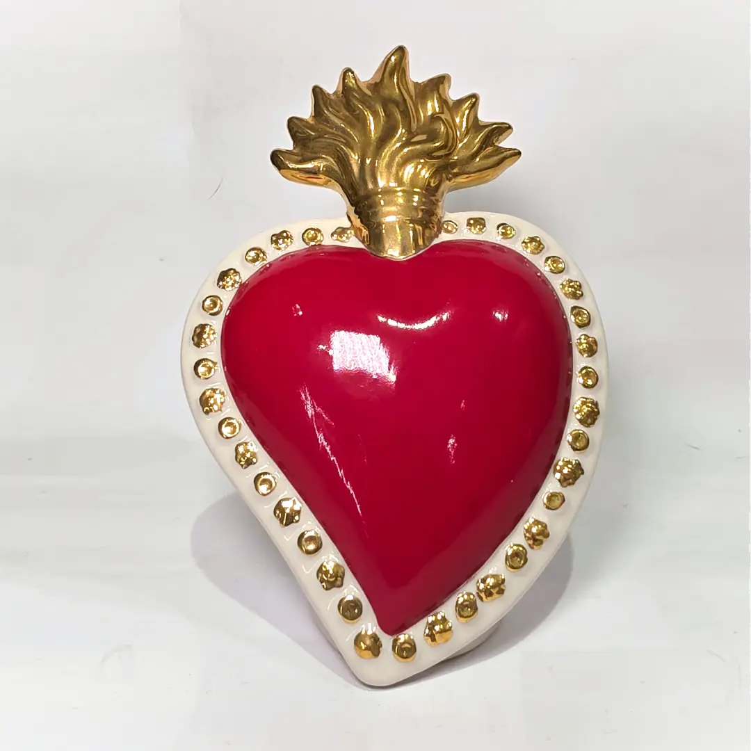 Scultura a forma di Cuore Sacro in ceramica rossa lucida, completata da una fiamma superiore dorata e una cornice bianca perlinata con borchie oro. Questo oggetto di design artigianale con base d'appoggio è un complemento d'arredo iconico, ideale per donare un tocco di eleganza e spiritualità moderna agli interni.