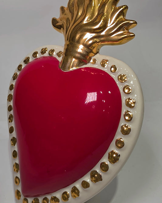 Scultura a forma di Cuore Sacro in ceramica rossa lucida, completata da una fiamma superiore dorata e una cornice bianca perlinata con borchie oro. Questo oggetto di design artigianale con base d'appoggio è un complemento d'arredo iconico, ideale per donare un tocco di eleganza e spiritualità moderna agli interni.