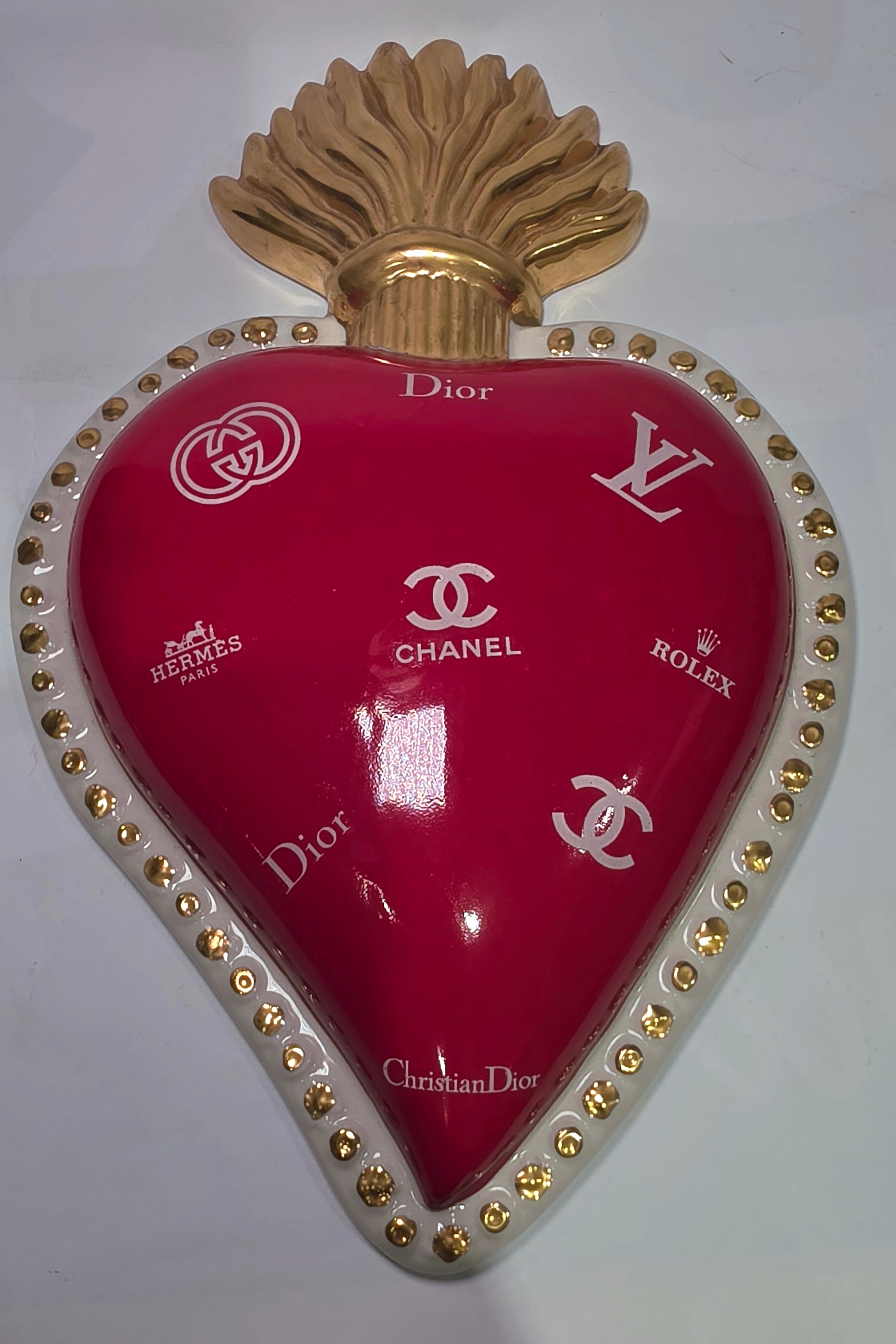 Scultura da parete a forma di Cuore Sacro grande in ceramica rossa lucida, decorata con diversi loghi fashion bianchi e sormontata da una fiamma dorata a rilievo. L'oggetto è rifinito con una cornice perlinata bianca e borchie oro, rappresentando un complemento d'arredo pop art dal design audace ideale per ambienti moderni.