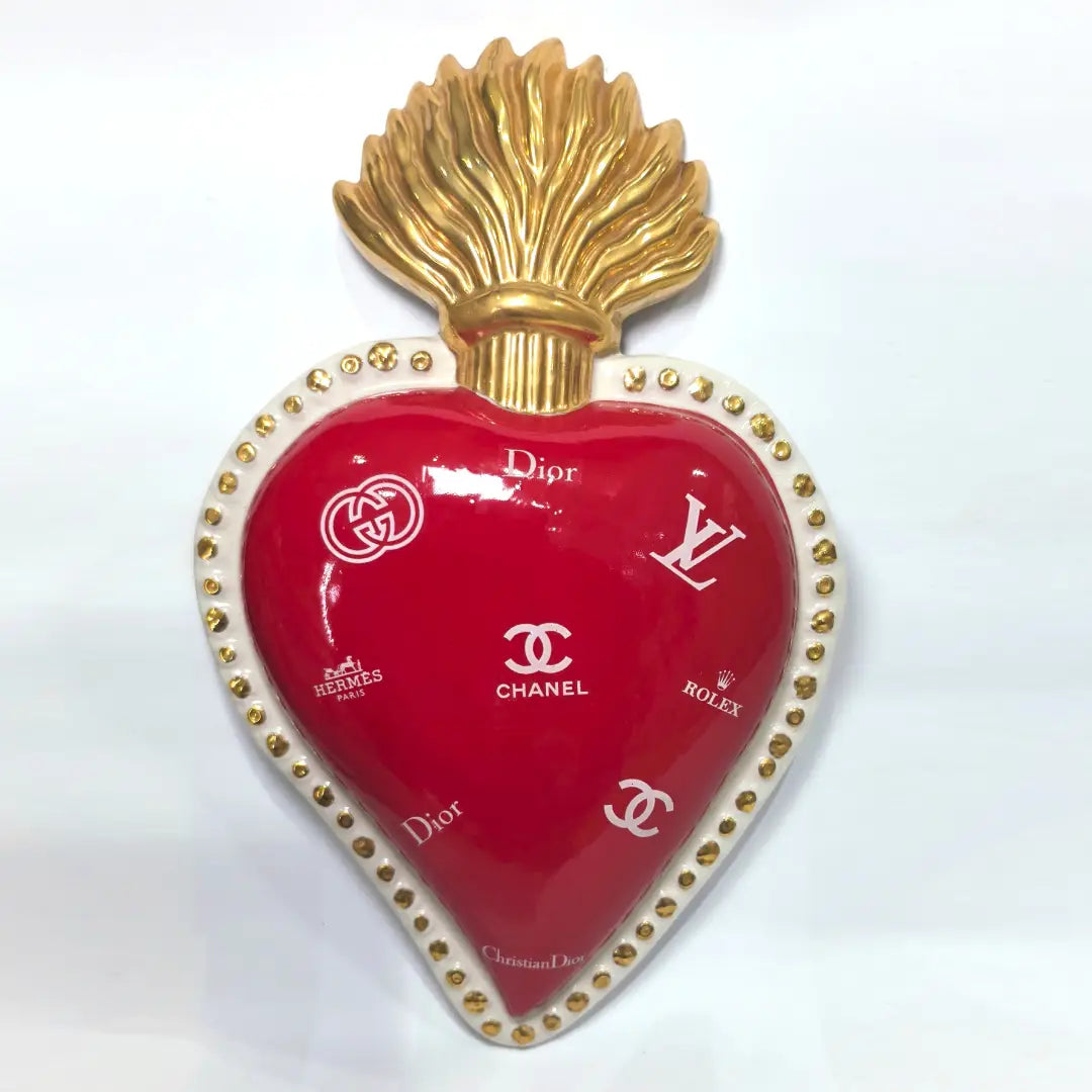 Scultura da parete a forma di Cuore Sacro grande in ceramica rossa lucida, decorata con diversi loghi fashion bianchi e sormontata da una fiamma dorata a rilievo. L'oggetto è rifinito con una cornice perlinata bianca e borchie oro, rappresentando un complemento d'arredo pop art dal design audace ideale per ambienti moderni.