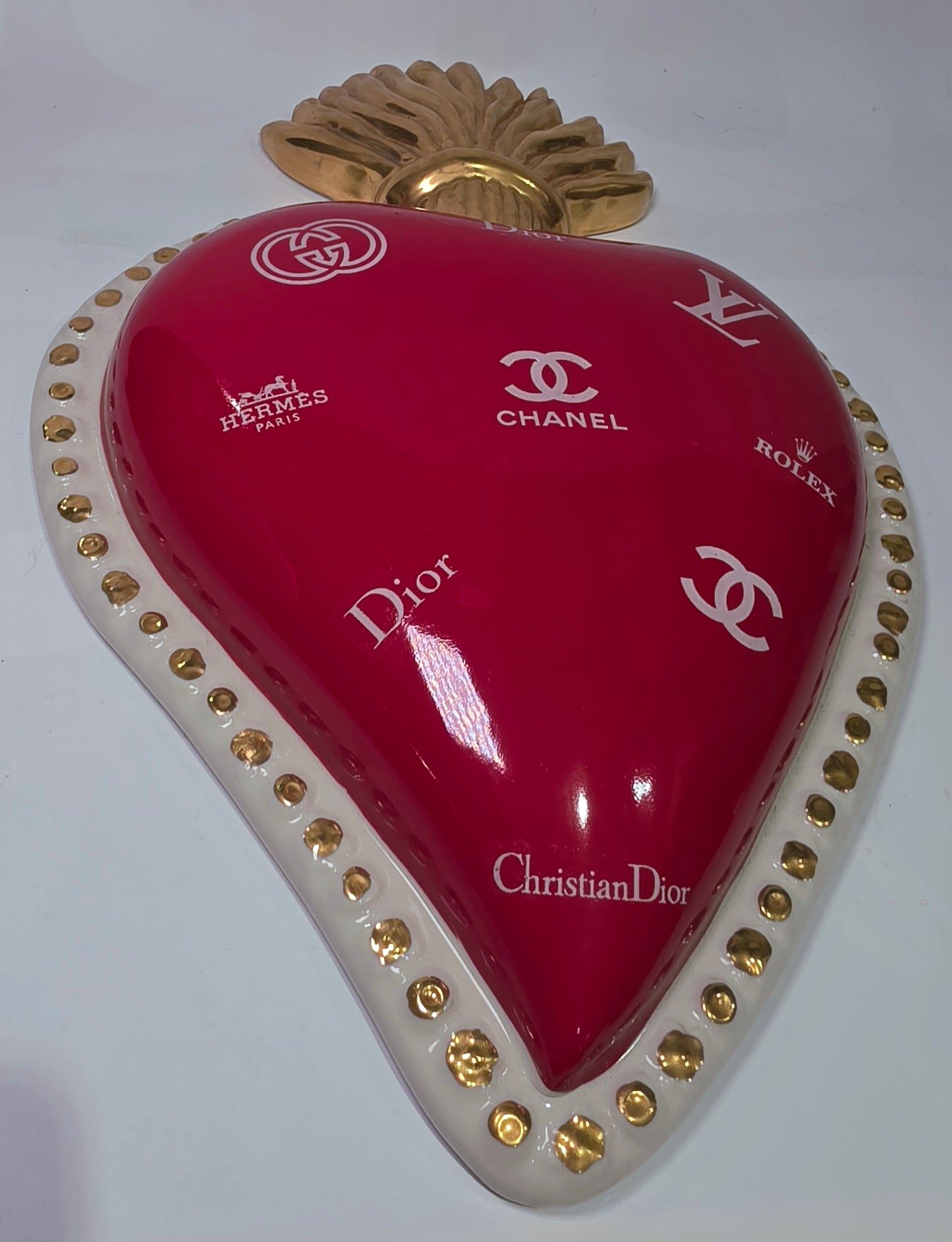 Scultura da parete a forma di Cuore Sacro grande in ceramica rossa lucida, decorata con diversi loghi fashion bianchi e sormontata da una fiamma dorata a rilievo. L'oggetto è rifinito con una cornice perlinata bianca e borchie oro, rappresentando un complemento d'arredo pop art dal design audace ideale per ambienti moderni.