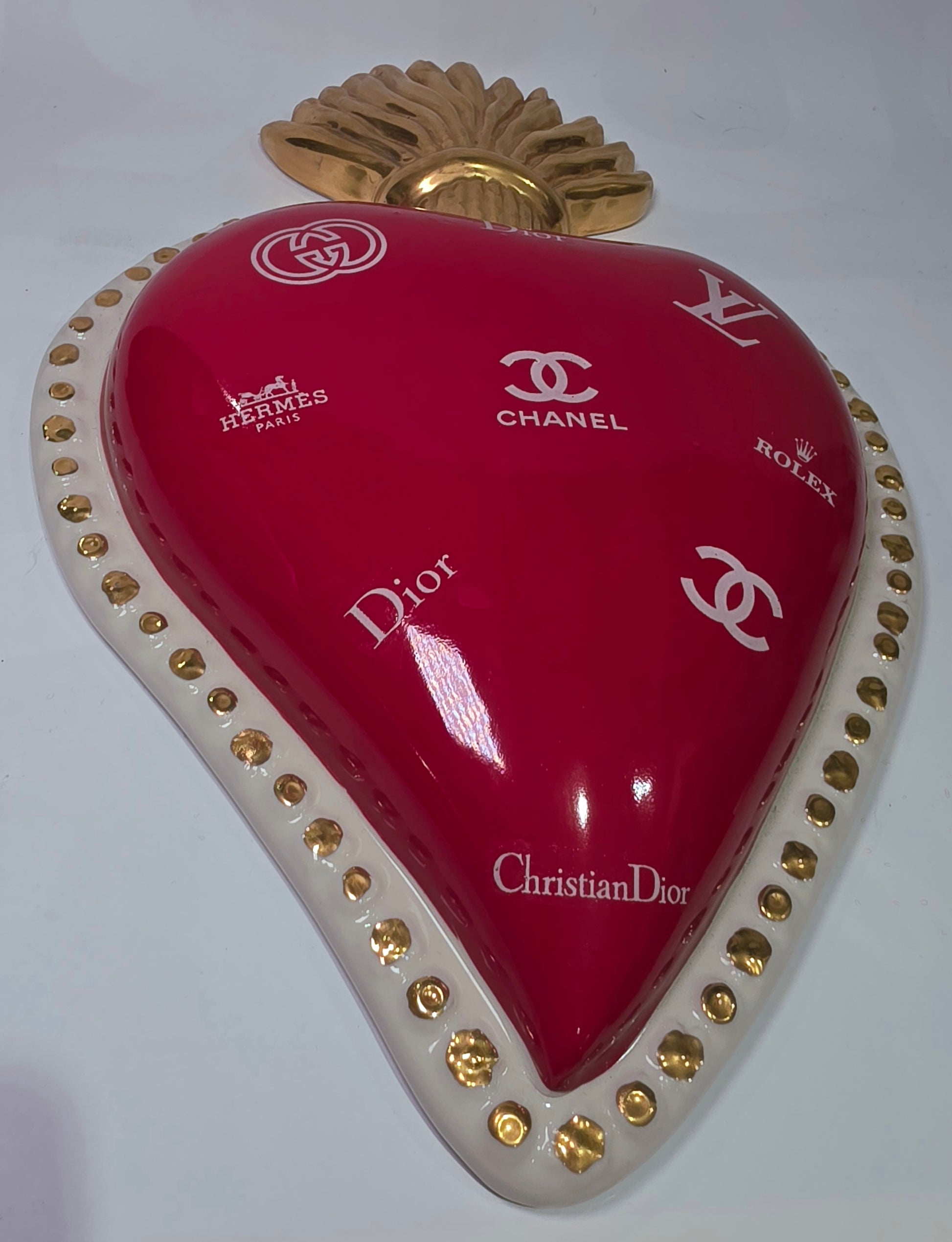 Scultura da parete a forma di Cuore Sacro grande in ceramica rossa lucida, decorata con diversi loghi fashion bianchi e sormontata da una fiamma dorata a rilievo. L'oggetto è rifinito con una cornice perlinata bianca e borchie oro, rappresentando un complemento d'arredo pop art dal design audace ideale per ambienti moderni.