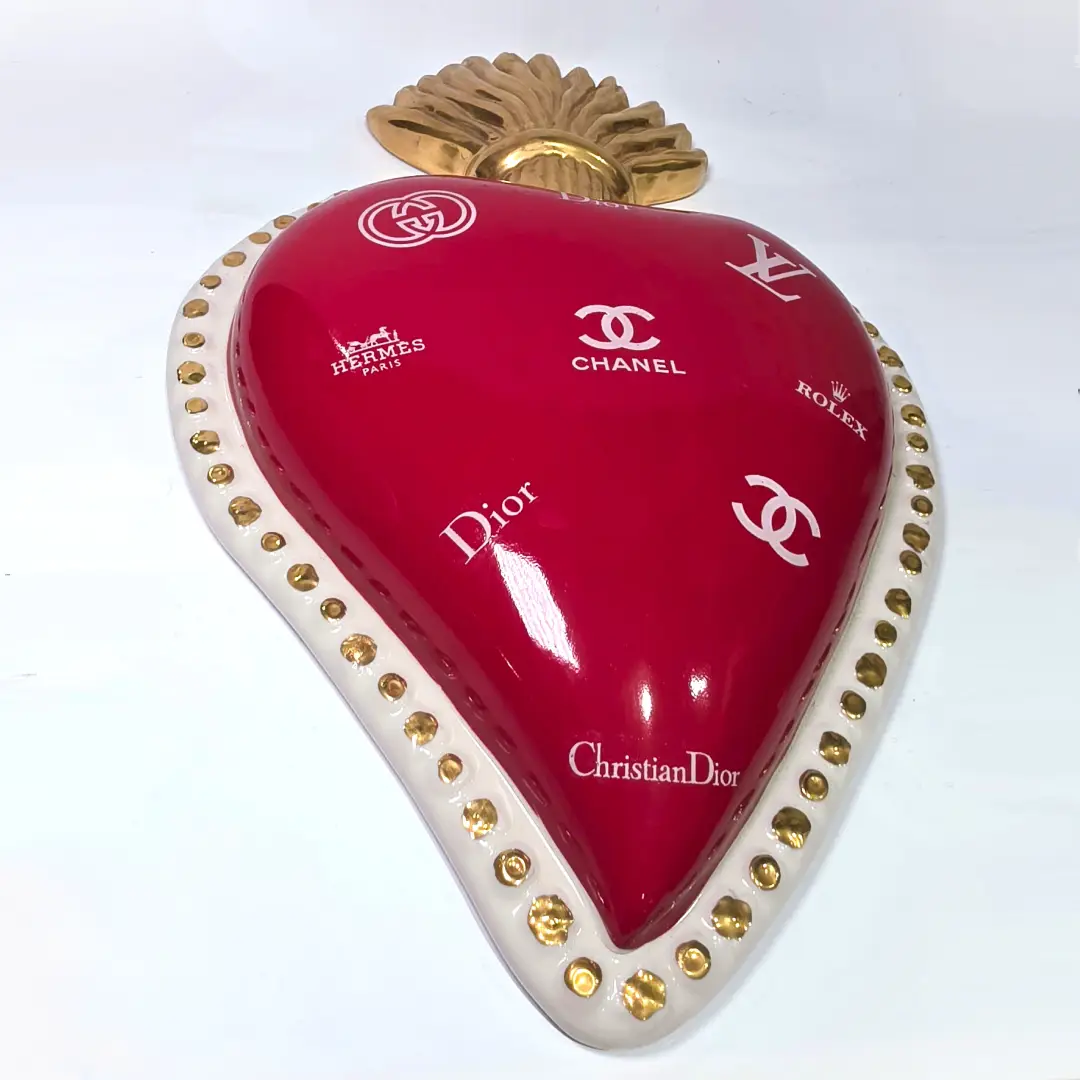 Scultura da parete a forma di Cuore Sacro grande in ceramica rossa lucida, decorata con diversi loghi fashion bianchi e sormontata da una fiamma dorata a rilievo. L'oggetto è rifinito con una cornice perlinata bianca e borchie oro, rappresentando un complemento d'arredo pop art dal design audace ideale per ambienti moderni.