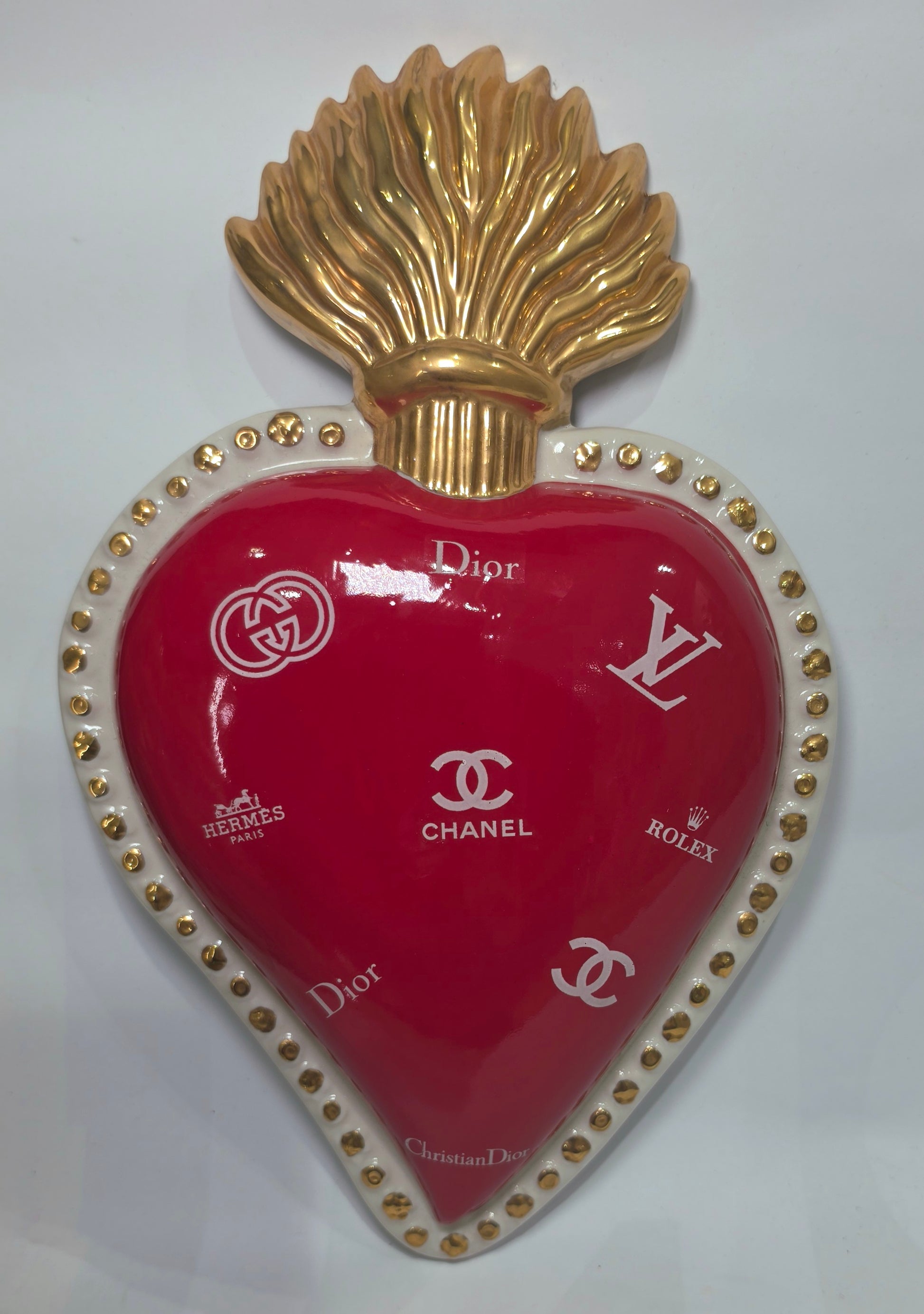 Scultura da parete a forma di Cuore Sacro grande in ceramica rossa lucida, decorata con diversi loghi fashion bianchi e sormontata da una fiamma dorata a rilievo. L'oggetto è rifinito con una cornice perlinata bianca e borchie oro, rappresentando un complemento d'arredo pop art dal design audace ideale per ambienti moderni.