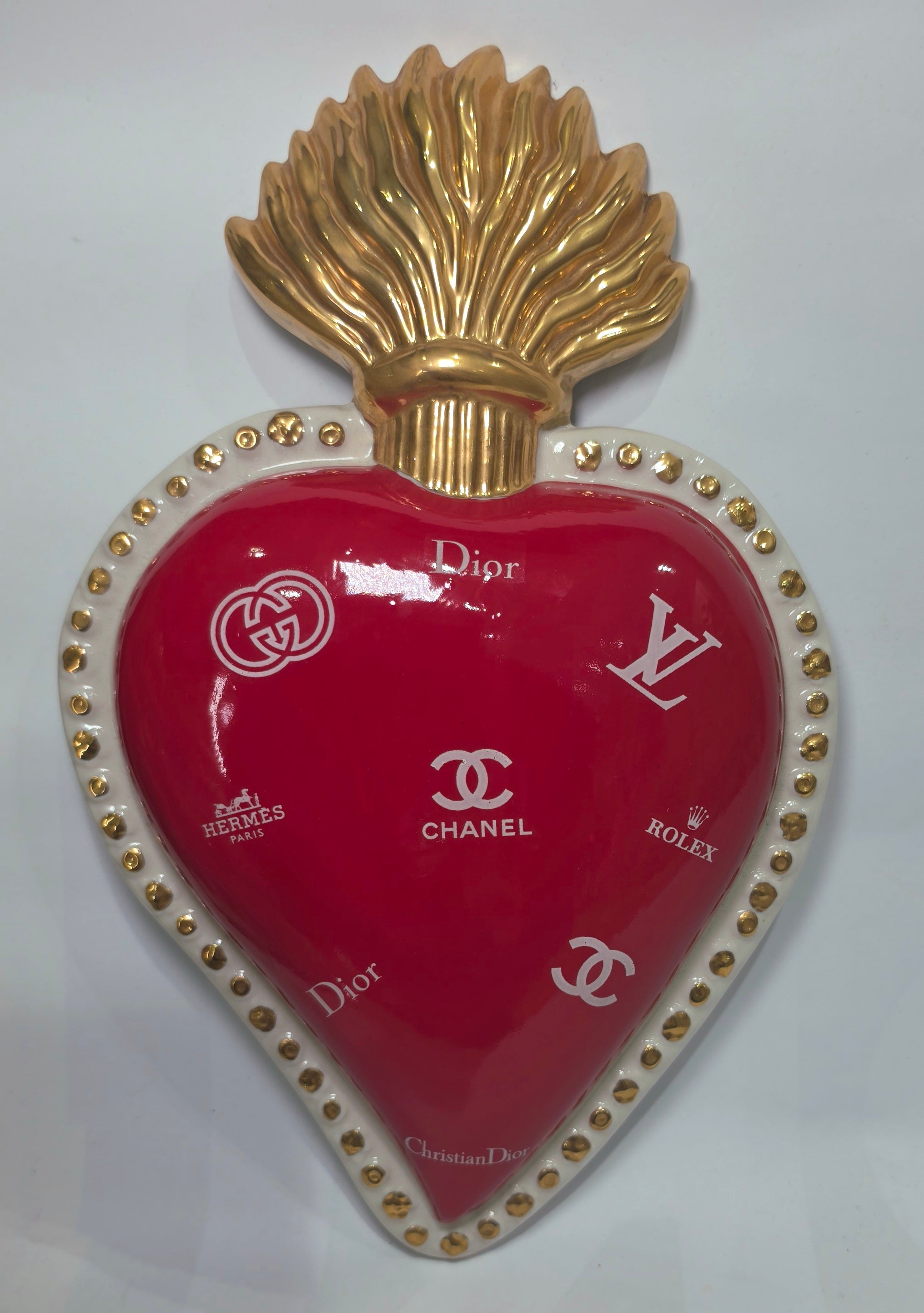 Scultura da parete a forma di Cuore Sacro grande in ceramica rossa lucida, decorata con diversi loghi fashion bianchi e sormontata da una fiamma dorata a rilievo. L'oggetto è rifinito con una cornice perlinata bianca e borchie oro, rappresentando un complemento d'arredo pop art dal design audace ideale per ambienti moderni.