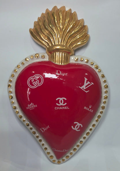 Scultura da parete a forma di Cuore Sacro grande in ceramica rossa lucida, decorata con diversi loghi fashion bianchi e sormontata da una fiamma dorata a rilievo. L'oggetto è rifinito con una cornice perlinata bianca e borchie oro, rappresentando un complemento d'arredo pop art dal design audace ideale per ambienti moderni.