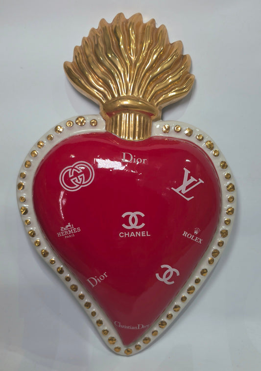 Scultura da parete a forma di Cuore Sacro grande in ceramica rossa lucida, decorata con diversi loghi fashion bianchi e sormontata da una fiamma dorata a rilievo. L'oggetto è rifinito con una cornice perlinata bianca e borchie oro, rappresentando un complemento d'arredo pop art dal design audace ideale per ambienti moderni.