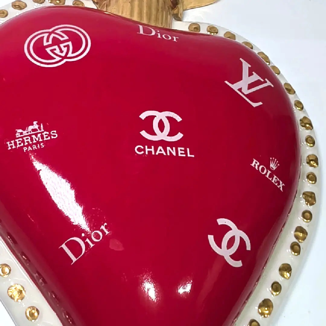 Scultura da parete a forma di Cuore Sacro grande in ceramica rossa lucida, decorata con diversi loghi fashion bianchi e sormontata da una fiamma dorata a rilievo. L'oggetto è rifinito con una cornice perlinata bianca e borchie oro, rappresentando un complemento d'arredo pop art dal design audace ideale per ambienti moderni.