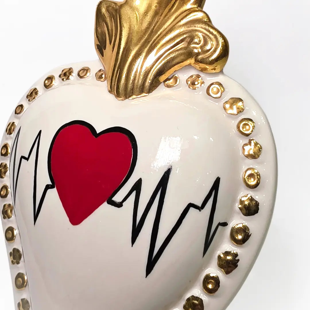 Scultura decorativa a forma di Cuore Sacro in ceramica bianca lucida, caratterizzata da un cuore rosso centrale sovrapposto a un disegno grafico nero che richiama il battito cardiaco. L'oggetto è impreziosito da una fiamma superiore dorata e una cornice perlinata con borchie oro, rendendolo un complemento d'arredo dal design moderno e originale.