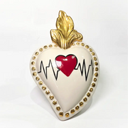 Scultura decorativa a forma di Cuore Sacro in ceramica bianca lucida, caratterizzata da un cuore rosso centrale sovrapposto a un disegno grafico nero che richiama il battito cardiaco. L'oggetto è impreziosito da una fiamma superiore dorata e una cornice perlinata con borchie oro, rendendolo un complemento d'arredo dal design moderno e originale.