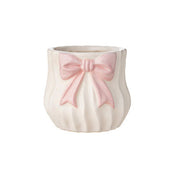 Vaso basso in ceramica bianca del marchio L'arte dei nacchi con design bombato e superficie decorata da scanalature verticali a effetto plissettato. Il prodotto presenta un elegante fiocco rosa applicato in rilievo sul fronte, caratterizzato da code lunghe e una finitura opaca, ideale come portaoggetti, piccolo vaso o bomboniera raffinata per eventi e decorazione d'interni.