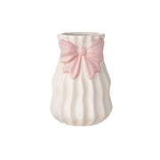 Vaso alto in ceramica bianca del marchio L'arte dei nacchi caratterizzato da una superficie con lavorazione a costine verticali ondulate che donano movimento al design. Il complemento d'arredo è impreziosito da un fiocco rosa decorativo in rilievo applicato sulla parte superiore vicino all'apertura, ideale come bomboniera o elemento decorativo per la casa dallo stile romantico e delicato.