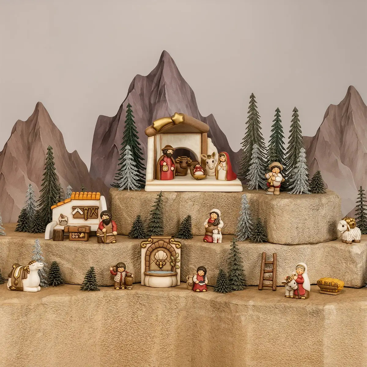 Presepe in ceramica ambientato su rocce a più livelli, con capanna centrale della Natività, casetta del falegname, fontana in pietra, pastori, pecore, cammello e alberi innevati sullo sfondo di montagne rocciose. Ambientazione completa dal design classico e curato firmata THUN, ideale come composizione natalizia o idea regalo per collezionisti.