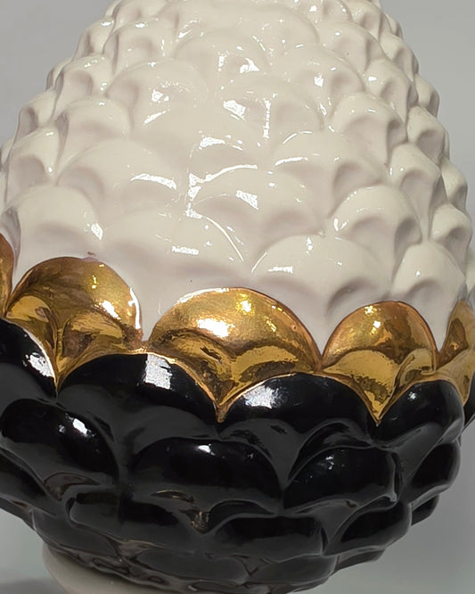 Pigna portafortuna in ceramica artigianale bicolore bianca e nera lucida, caratterizzata da una fascia centrale dorata che separa le squame in rilievo. Questo elegante oggetto ornamentale con base d'appoggio rifinita in oro è un complemento d'arredo di design ideale per donare un tocco di prestigio e tradizione siciliana agli ambienti contemporanei.