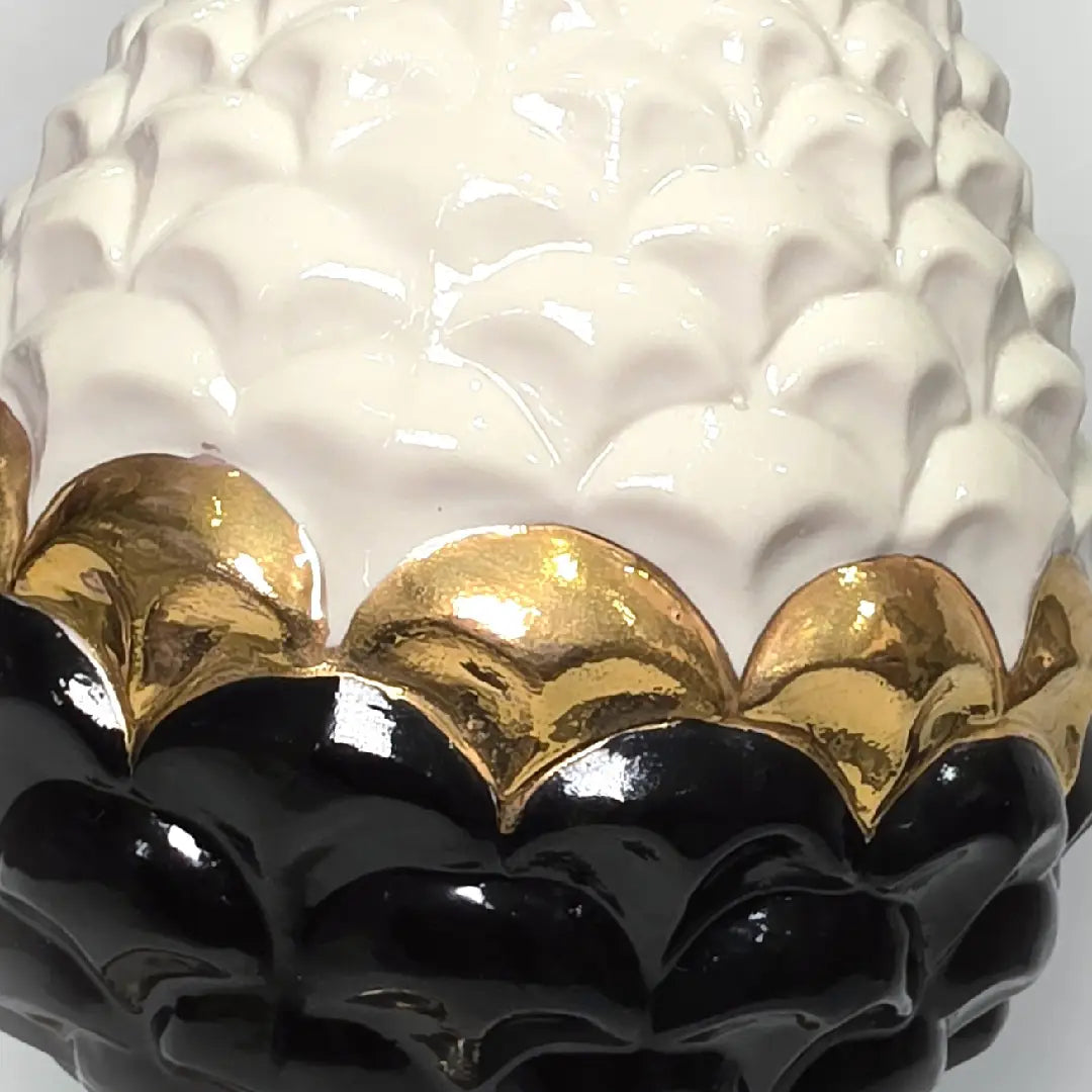 Pigna portafortuna in ceramica artigianale bicolore bianca e nera lucida, caratterizzata da una fascia centrale dorata che separa le squame in rilievo. Questo elegante oggetto ornamentale con base d'appoggio rifinita in oro è un complemento d'arredo di design ideale per donare un tocco di prestigio e tradizione siciliana agli ambienti contemporanei.