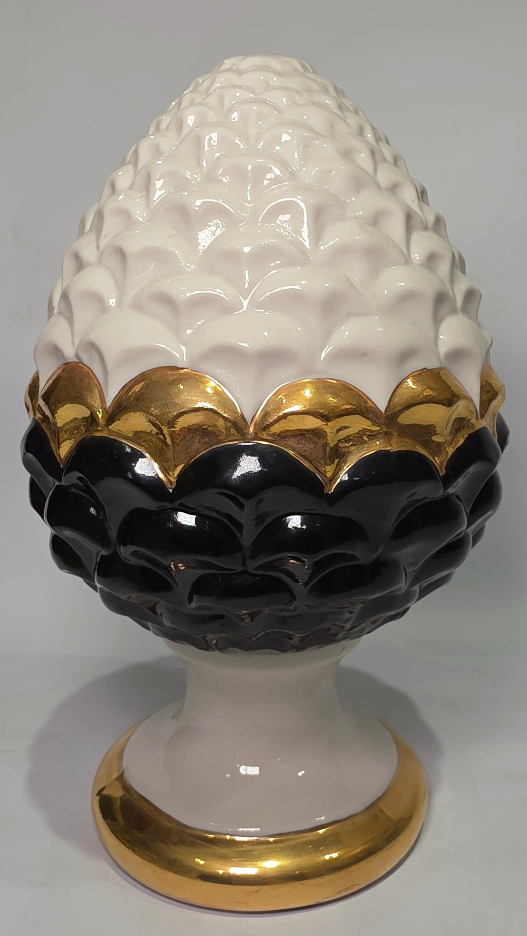 Pigna portafortuna in ceramica artigianale bicolore bianca e nera lucida, caratterizzata da una fascia centrale dorata che separa le squame in rilievo. Questo elegante oggetto ornamentale con base d'appoggio rifinita in oro è un complemento d'arredo di design ideale per donare un tocco di prestigio e tradizione siciliana agli ambienti contemporanei.