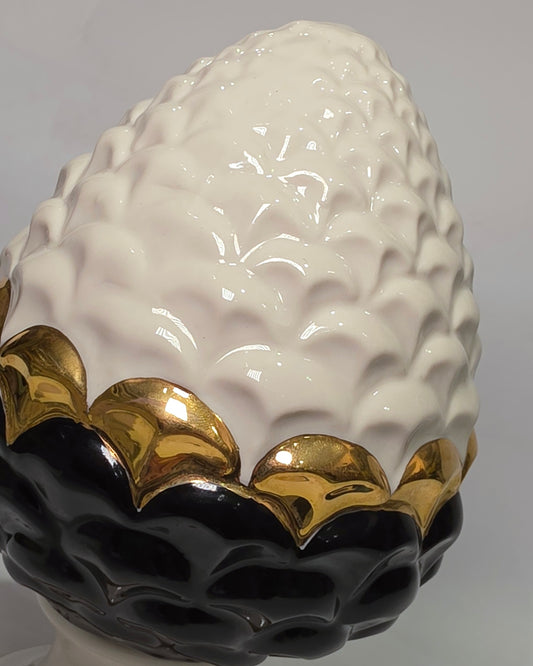 Pigna portafortuna in ceramica artigianale bicolore bianca e nera lucida, caratterizzata da una fascia centrale dorata che separa le squame in rilievo. Questo elegante oggetto ornamentale con base d'appoggio rifinita in oro è un complemento d'arredo di design ideale per donare un tocco di prestigio e tradizione siciliana agli ambienti contemporanei.