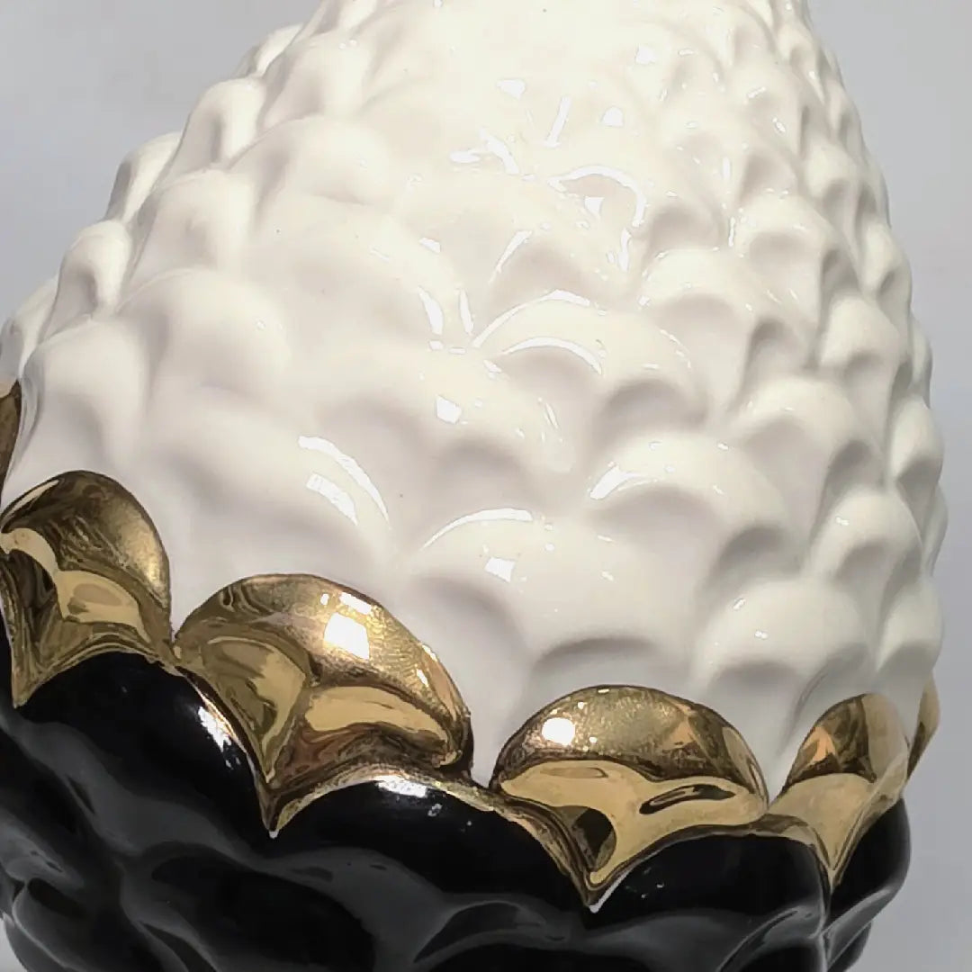 Pigna portafortuna in ceramica artigianale bicolore bianca e nera lucida, caratterizzata da una fascia centrale dorata che separa le squame in rilievo. Questo elegante oggetto ornamentale con base d'appoggio rifinita in oro è un complemento d'arredo di design ideale per donare un tocco di prestigio e tradizione siciliana agli ambienti contemporanei.
