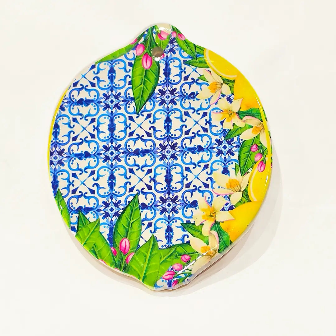 Sottobicchiere in ceramica a forma di limone, disponibile in due varianti decorative: una gialla lucida con foglie verdi e boccioli rosa, l'altra con fantasia maiolica blu e bianca abbinata a fiori di zagara. Set assortito di sottobicchieri artigianali a tema mediterraneo, perfetto per la tavola estiva o come idea regalo souvenir.