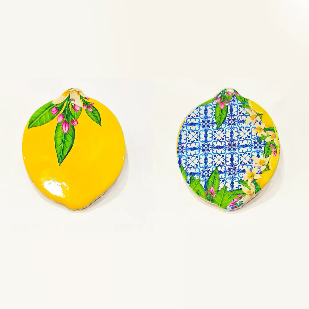 Sottobicchiere in ceramica a forma di limone, disponibile in due varianti decorative: una gialla lucida con foglie verdi e boccioli rosa, l'altra con fantasia maiolica blu e bianca abbinata a fiori di zagara. Set assortito di sottobicchieri artigianali a tema mediterraneo, perfetto per la tavola estiva o come idea regalo souvenir.