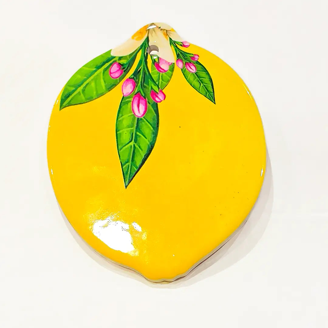 Sottobicchiere in ceramica a forma di limone, disponibile in due varianti decorative: una gialla lucida con foglie verdi e boccioli rosa, l'altra con fantasia maiolica blu e bianca abbinata a fiori di zagara. Set assortito di sottobicchieri artigianali a tema mediterraneo, perfetto per la tavola estiva o come idea regalo souvenir.