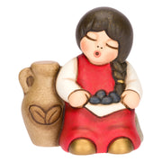 Statuina in ceramica raffigurante una bimba del presepe con treccia nera e vestito rosso e bianco, seduta accanto a un'anfora mentre raccoglie olive nel grembo. Personaggio dal design classico e curato firmato THUN, ideale per arricchire il presepe o come idea regalo natalizia.