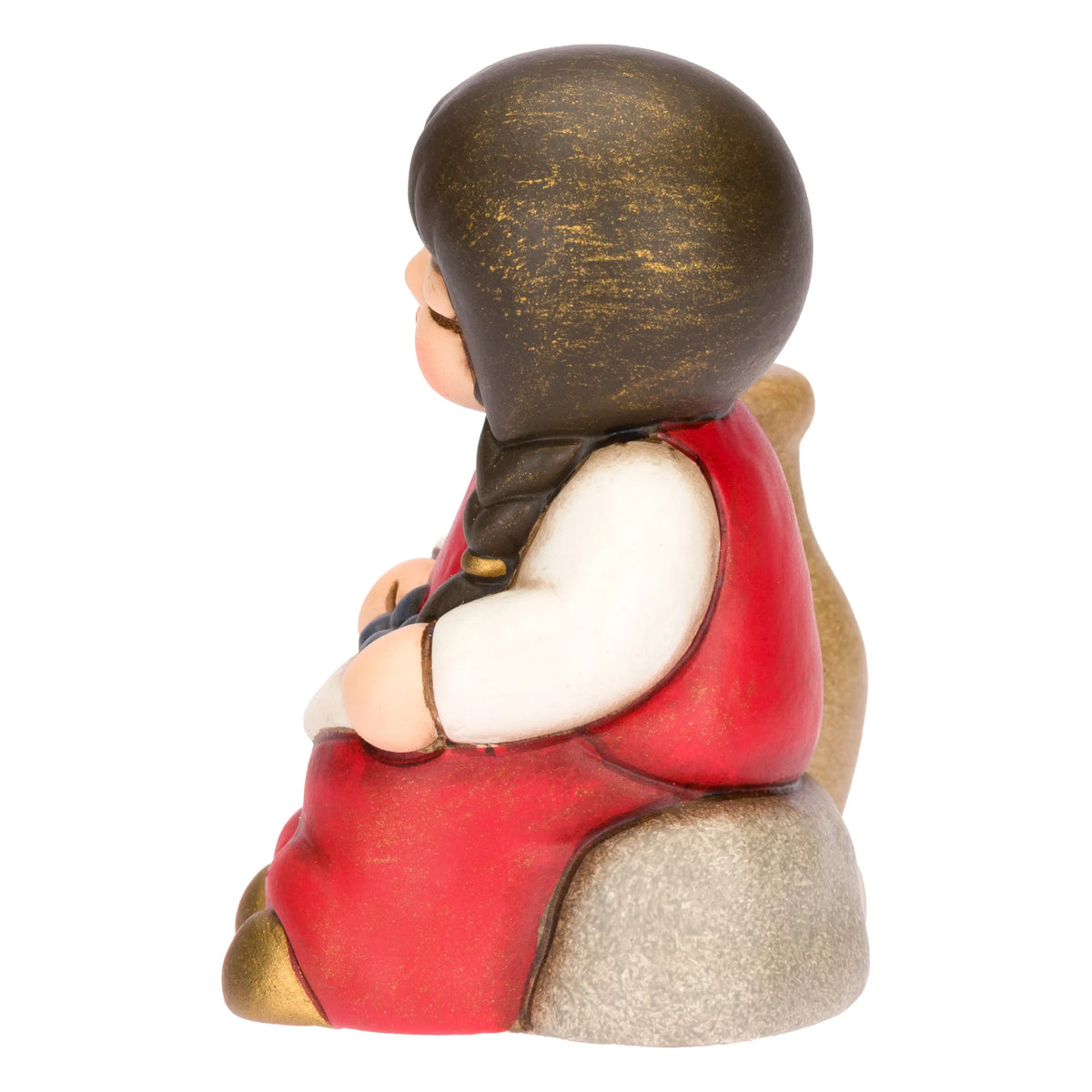 Statuina in ceramica raffigurante una bimba del presepe con treccia nera e vestito rosso e bianco, seduta accanto a un'anfora mentre raccoglie olive nel grembo. Personaggio dal design classico e curato firmato THUN, ideale per arricchire il presepe o come idea regalo natalizia.