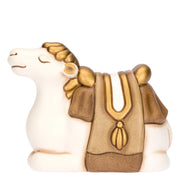 Statuina in ceramica raffigurante un cammello del presepe seduto, con manto bianco e gobba coperta da un ricco basto marrone e oro decorato con nappe. Personaggio dal design classico e curato firmato THUN, ideale per arricchire il presepe o come idea regalo natalizia.