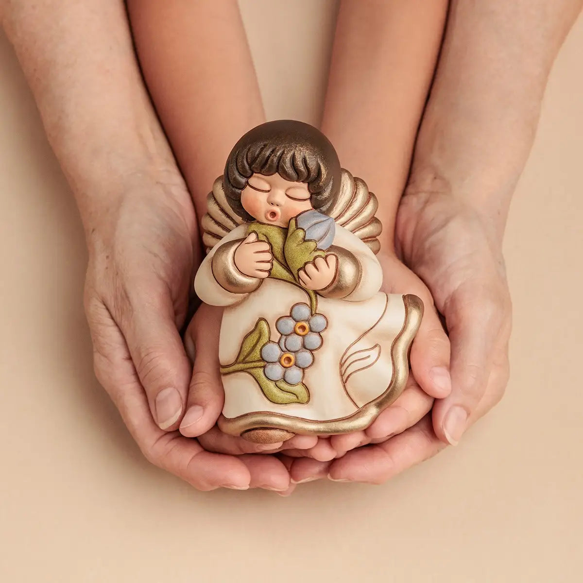 Statuina in ceramica Thun dell'Angelo dei Nonni Anna, collezione 2025. L'angelo è raffigurato con gli occhi chiusi, ali dorate e capelli castani. Indossa una veste color crema con bordi dorati e decorazioni floreali azzurre sulla parte inferiore. L'angelo tiene tra le mani un mazzo di fiori azzurri con foglie verdi, portandoli vicino al viso. Design classico Thun con finitura lucida e forme arrotondate.