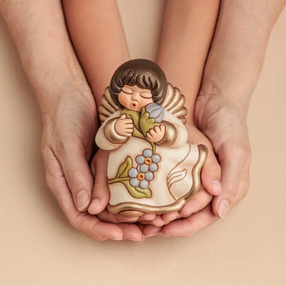 Statuina in ceramica Thun dell'Angelo dei Nonni Anna, collezione 2025. L'angelo è raffigurato con gli occhi chiusi, ali dorate e capelli castani. Indossa una veste color crema con bordi dorati e decorazioni floreali azzurre sulla parte inferiore. L'angelo tiene tra le mani un mazzo di fiori azzurri con foglie verdi, portandoli vicino al viso. Design classico Thun con finitura lucida e forme arrotondate.