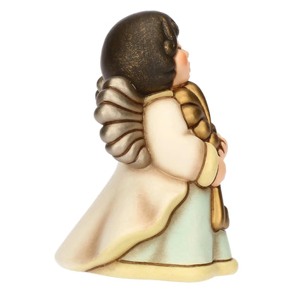Statuina in ceramica che raffigura un angelo di profilo, caratterizzata da forme morbide e toni caldi e sfumati. L'angelo ha i capelli scuri corti, ali dorate con piume stilizzate e arrotondate, e indossa una tunica chiara composta da un mantello bianco panna sovrapposto a una veste color acquamarina tenue. I bordi della tunica sono rifiniti in giallo pallido. L'angelo tiene tra le mani un oggetto dorato e voluminoso, mentre il volto è rivolto leggermente verso l'alto con un'espressione serena.