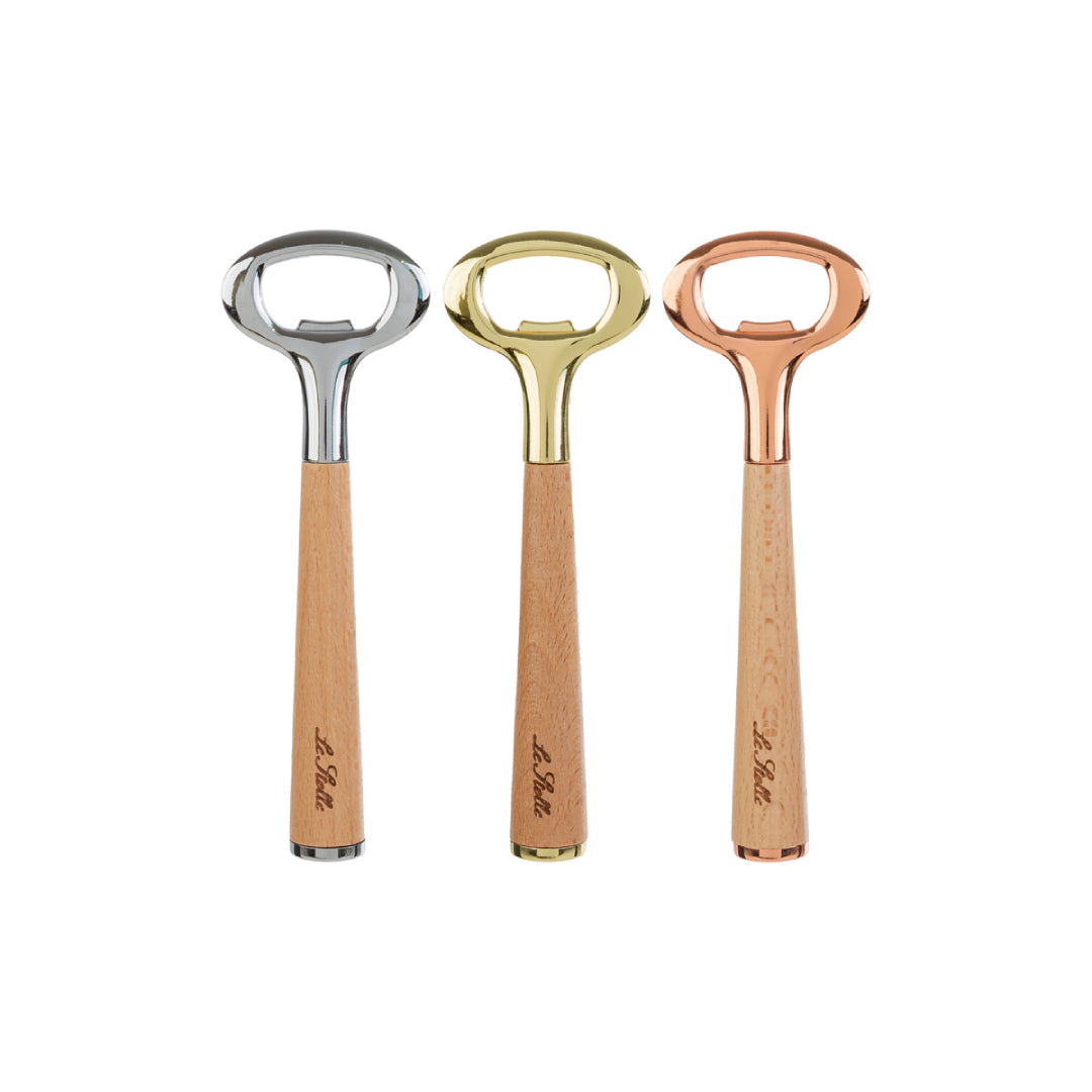 Apribottiglie con manico in legno chiaro inciso "Le Stelle" e testa in metallo, disponibile in tre finiture assortite argento, oro e rosé, presentato accanto alla sua confezione bianca con logo del marchio. Utensile da cucina elegante e funzionale, ideale come bomboniera o idea regalo raffinata.
