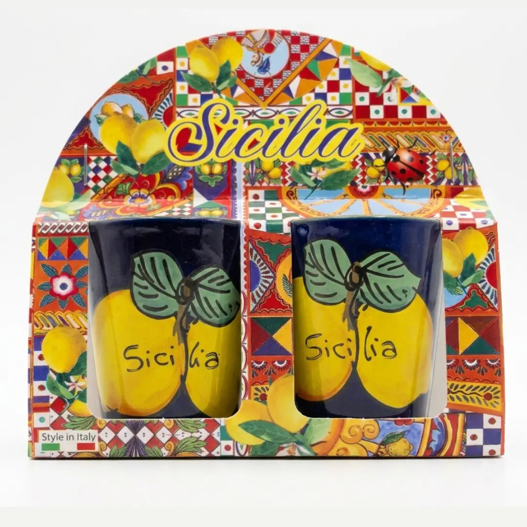 Coppia di bicchierini in ceramica decorati con vivaci limoni e la scritta "Sicilia" su sfondo blu scuro o giallo brillante, perfetti per servire liquori come il limoncello. Questa coppia di tazzine artigianali si presenta in una confezione regalo assortita in stile maiolica siciliana, ideale come souvenir colorato o come bomboniera per eventi speciali.