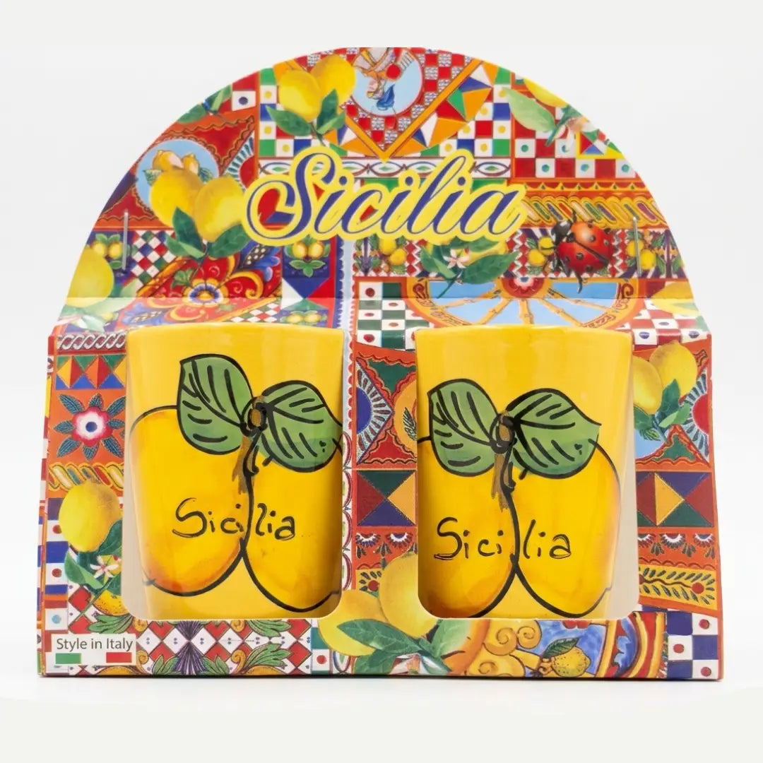 Coppia di bicchierini in ceramica decorati con vivaci limoni e la scritta "Sicilia" su sfondo blu scuro o giallo brillante, perfetti per servire liquori come il limoncello. Questa coppia di tazzine artigianali si presenta in una confezione regalo assortita in stile maiolica siciliana, ideale come souvenir colorato o come bomboniera per eventi speciali.