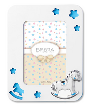 Portafoto per bambina in legno bianco con cornice sagomata e angoli arrotondati. Presenta trafori a forma di stelle e di un cavallino a dondolo che rivelano uno sfondo celeste pastello. In basso a destra è applicato un cavallino a dondolo in argento bilaminato con dettagli rosa sulle zampe e sulla sella. L'interno mostra un inserto decorativo con stelle e il logo "Brera - Real Italian Style".