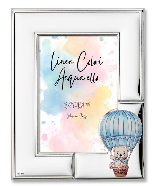 Portafoto da tavolo per bambino con cornice in argento bilaminato lucido. L'angolo in basso a destra è decorato con un'applicazione smaltata in rilievo che raffigura un dolce orsetto seduto nel cestino di una mongolfiera a strisce celeste e panna. All'interno è presente l'inserto "Linea Colori Acquarello - Brera 216 - Made in Italy" su sfondo sfumato pastello.