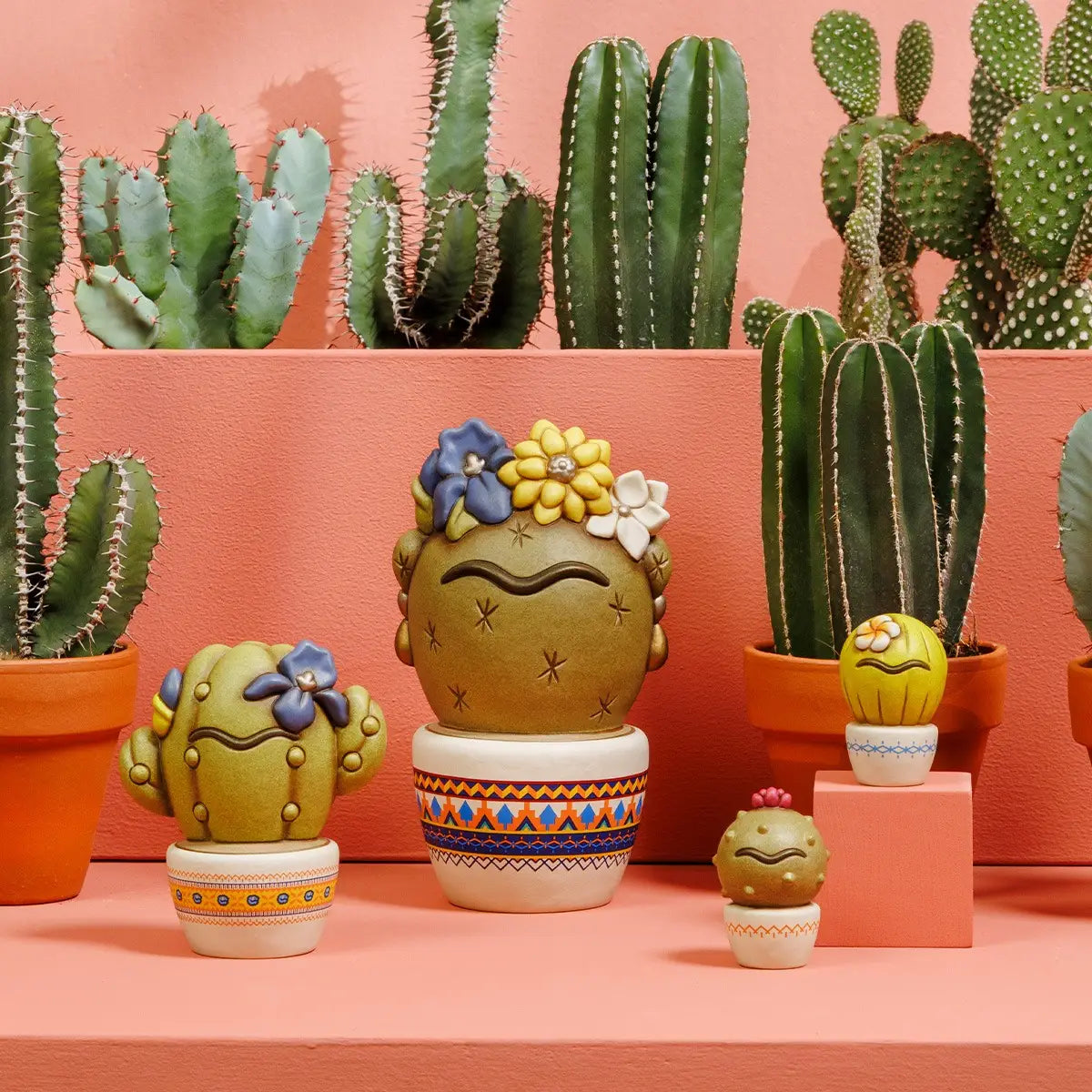 Set di statuine in ceramica Thun della collezione Cactus Frida, con diverse varianti di piante grasse stilizzate ornate da fiori colorati e dettagli ispirati a Frida Kahlo. Questi complementi d'arredo creativi, esposti in un ambientazione con veri cactus, sono ideali per aggiungere un tocco artistico e gioioso a qualsiasi angolo della casa.