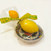 Calamita con sapone profumato al limone "Amalfi", a forma di limone giallo lucido, confezionata su un piattino in ceramica siciliana dipinta a mano con motivi floreali blu e arancio, con fiocco in raso giallo a stampa di limoni. Articolo souvenir della costiera amalfitana, ideale come bomboniera o idea regalo, in confezione trasparente con piccola saponetta arancione a forma di frutto abbinata.