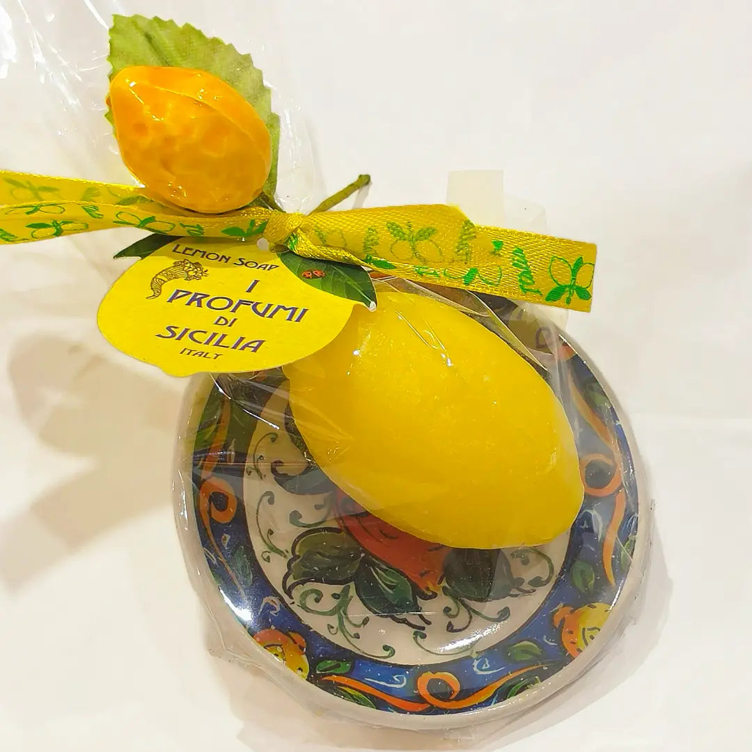 Calamita con sapone profumato al limone "Amalfi", a forma di limone giallo lucido, confezionata su un piattino in ceramica siciliana dipinta a mano con motivi floreali blu e arancio, con fiocco in raso giallo a stampa di limoni. Articolo souvenir della costiera amalfitana, ideale come bomboniera o idea regalo, in confezione trasparente con piccola saponetta arancione a forma di frutto abbinata.