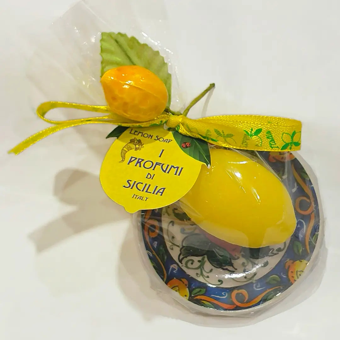 Calamita con sapone profumato al limone "Amalfi", a forma di limone giallo lucido, confezionata su un piattino in ceramica siciliana dipinta a mano con motivi floreali blu e arancio, con fiocco in raso giallo a stampa di limoni. Articolo souvenir della costiera amalfitana, ideale come bomboniera o idea regalo, in confezione trasparente con piccola saponetta arancione a forma di frutto abbinata.