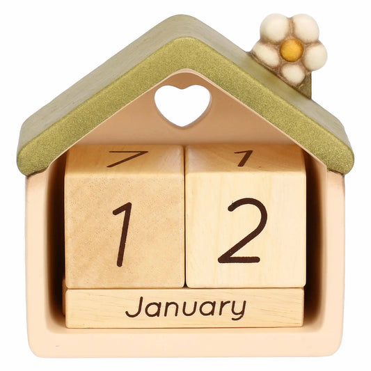 Un calendario perpetuo da tavolo in ceramica e legno, modellato a forma di piccola casa stilizzata. La struttura esterna è color crema con un tetto spiovente verde salvia, decorato sulla sommità da un piccolo fiore bianco con il centro giallo e caratterizzato da un foro a forma di cuore sulla facciata anteriore. All'interno della cavità della casetta sono alloggiati tre elementi mobili in legno chiaro: due cubi superiori che mostrano le cifre dei giorni e un blocchetto con il mese