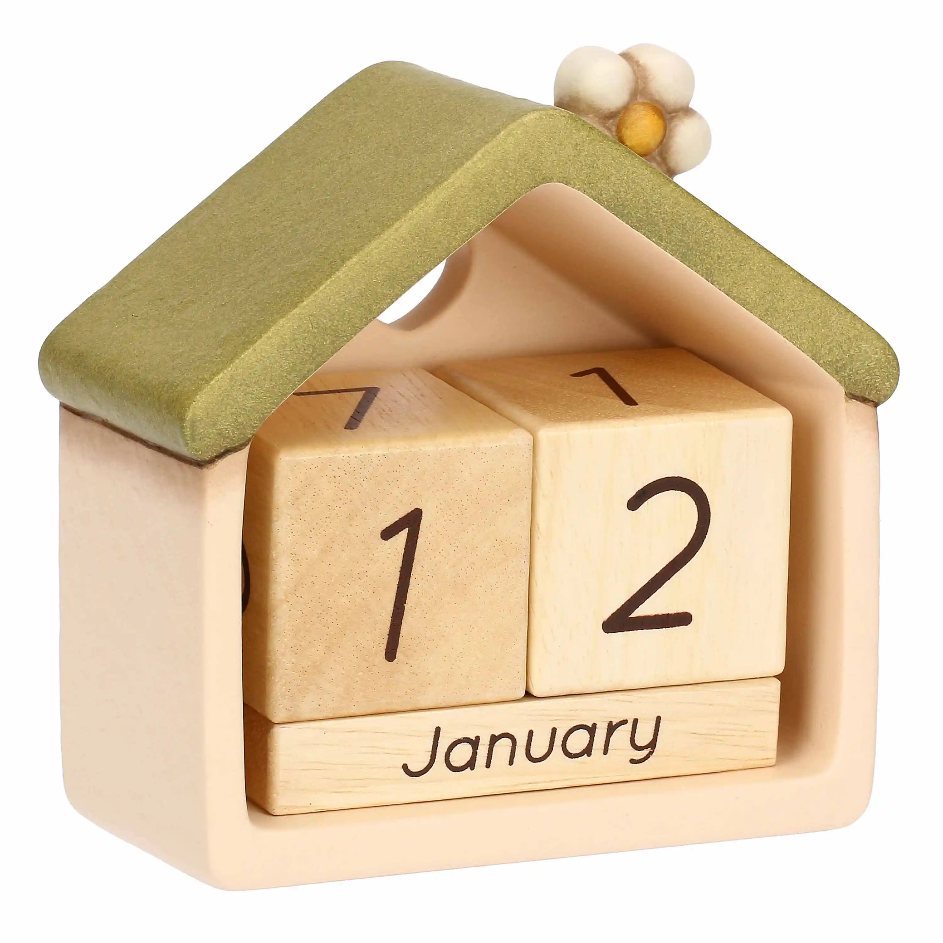 Una prospettiva angolata del calendario che ne evidenzia la profondità e la struttura tridimensionale. La casetta in ceramica color crema ospita al suo interno i due cubi numerici in legno e il blocchetto dei mesi leggermente rientrati rispetto al bordo frontale. Il tetto verde salvia e il fiorellino decorativo sono visti lateralmente, mostrando la consistenza opaca del materiale, mentre il foro a cuore crea un gioco di luce attraverso lo schienale della struttura.