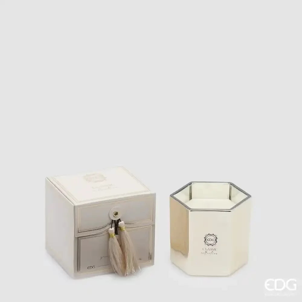 Candela profumata Classic dorata alla fragranza Oud firmata EDG Enzo de Gasperi, custodita in un raffinato bicchiere esagonale in vetro con finitura lucida a specchio. Questa candela decorativa di lusso è affiancata dalla sua elegante confezione regalo a scatola nei toni del bianco e dell'oro, impreziosita da una chiusura con nappa in tessuto coordinata.