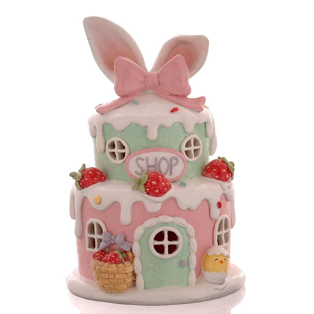 Decorazione pasquale scultorea Vetur in poliresina con luce LED. Casetta dei dolci a due piani in rosa e verde pastello, decorata con fragole, glassa bianca effetto neve, orecchie da coniglio giganti e insegna "Shop".