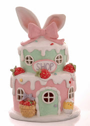 Decorazione pasquale scultorea Vetur in poliresina con luce LED. Casetta dei dolci a due piani in rosa e verde pastello, decorata con fragole, glassa bianca effetto neve, orecchie da coniglio giganti e insegna "Shop".