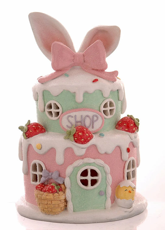 Decorazione pasquale scultorea Vetur in poliresina con luce LED. Casetta dei dolci a due piani in rosa e verde pastello, decorata con fragole, glassa bianca effetto neve, orecchie da coniglio giganti e insegna "Shop".