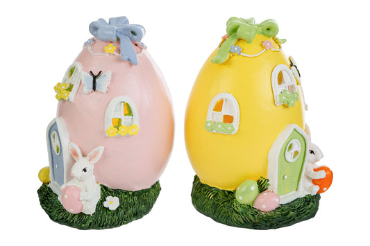 Coppia di decorazioni pasquali a forma di casa-uovo in resina con illuminazione interna a LED. Una versione è rosa con porta azzurra e l'altra gialla con porta verde. Entrambe presentano dettagli di finestrelle fiorite, farfalle applicate, un fiocco sulla sommità e un coniglietto bianco alla base con uova colorate.