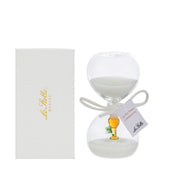 Clessidra in vetro trasparente riempita di sabbia bianca, con un calice dorato e un rametto verde all'interno del bulbo inferiore e fiocco bianco con etichetta "La mia prima Comunione". Bomboniera decorativa presentata con la sua confezione bianca del marchio Le Stelle, ideale come ricordo per la Prima Comunione.