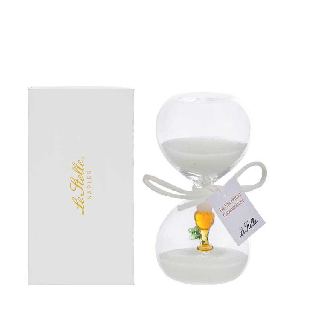 Clessidra in vetro trasparente riempita di sabbia bianca, con un calice dorato e un rametto verde all'interno del bulbo inferiore e fiocco bianco con etichetta "La mia prima Comunione". Bomboniera decorativa presentata con la sua confezione bianca del marchio Le Stelle, ideale come ricordo per la Prima Comunione.